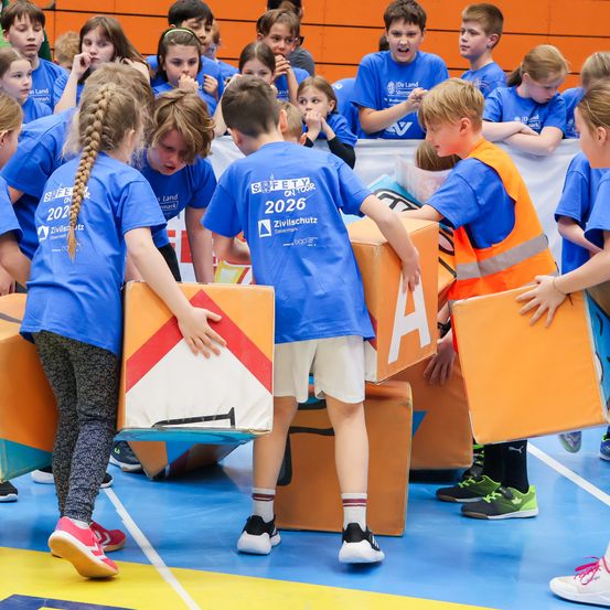 Kinder in blauen T-Shirts stehen um orangefarbene Kisten herum, eines trägt eine Sicherheitsweste. Sie scheinen an einer Veranstaltung oder Aktivität auf einem Turnhallenboden teilzunehmen.