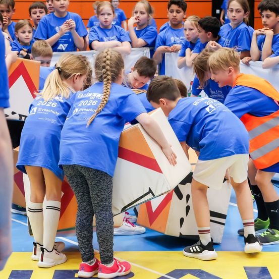 Schulkinder in blauen Shirts spielen auf einer bunten Matte in einer Turnhalle. Einige tragen Turnschuhe, andere reflektierende Westen. Eine Gruppe beobachtet von hinten.