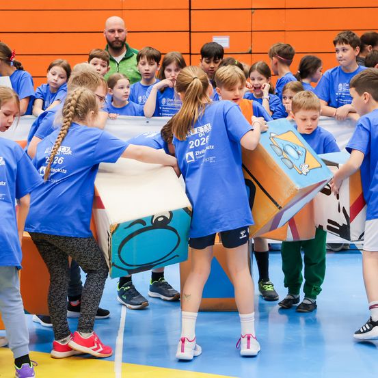 Kinder in blauen T-Shirts mit Nummern darauf spielen mit Kisten auf einem Turnhallenboden. Sie sind von anderen Kindern und einem erwachsenen Mann, der zusieht, umgeben.