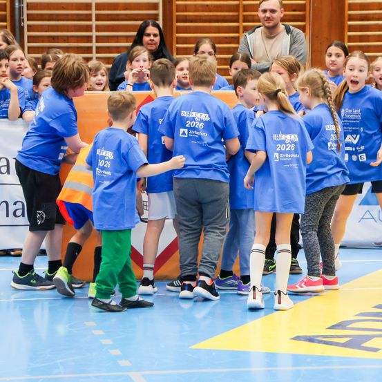 Eine Gruppe von Kindern in blauen Trikots mit der Nummer 2026 versammelt sich in einer Sporthalle, einige jubeln. Dahinter steht ein Erwachsener und beobachtet.