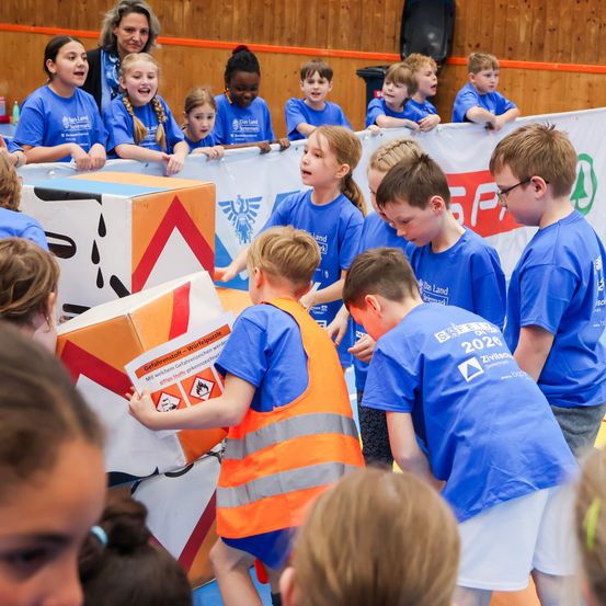 Kinder in blauen T-Shirts versammeln sich um eine große, bunte Kiste mit der Aufschrift 'Gefahrenstoff-Wurfpuzzle'. Sie sind in einer Aktivität verstrickt, wobei sich einige über die Kiste lehnen. Dahinter wird ein Banner mit einem Logo und Text gezeigt.