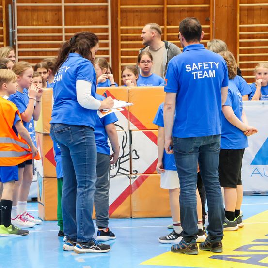 Eine Gruppe von Kindern und Erwachsenen ist in einer Turnhalle versammelt. Einige tragen blaue Uniformen, andere orange Weste. Eine Frau hält eine Clipboard in der Hand, wahrscheinlich spricht sie die Gruppe an. Auf einem Mann steht Sicherheitsteam.