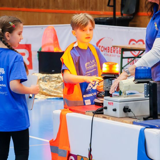 Ein junger Junge in einer Sicherheitsweste interagiert mit einer Maschine bei einer Schulveranstaltung, während ein Mädchen in einem blauen Shirt zusieht. Eine Frau beaufsichtigt die Aktivität.