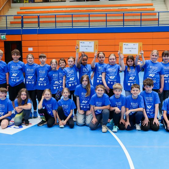 Eine Gruppe von Kindern in passenden blauen T-Shirts mit dem Text 'AV' posiert für ein Foto auf einem blauen Boden. Sie halten Schilder mit Namen und Nummern. Dahinter ist ein unscharfer Hintergrund eines Fitnessstudios.
