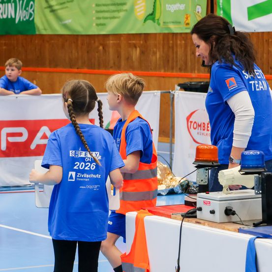 Drei Kinder in blauen und orangefarbenen Sicherheitswesten stehen neben einer Frau in einem blauen Shirt. Sie befinden sich in einer Turnhalle mit einem Sicherheitsteam-Banner und anderen Schildern im Hintergrund.