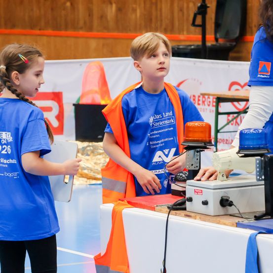 Zwei Kinder in blauen T-Shirts stehen neben einem Tisch. Eines hält eine Clipboard und das andere steht vor einem Gerät. Hinter ihnen ist eine Holzwand.