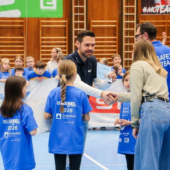 Ein Mann überreicht zwei Mädchen in einer Turnhalle ein Zertifikat vor einer Menge Kinder und Erwachsener.