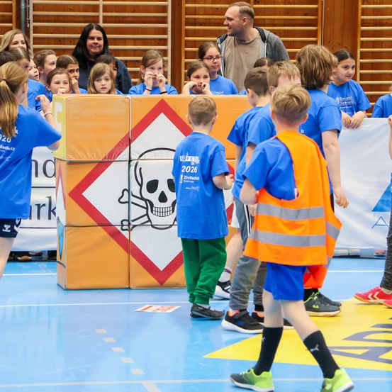 Eine Gruppe von Kindern in blauen T-Shirts versammelt sich um eine Kiste mit Totenkopf- und Knochenkreuz-Etikett. Sie befinden sich auf einem Hallenboden, mit Erwachsenen und weiteren Kindern hinter ihnen.