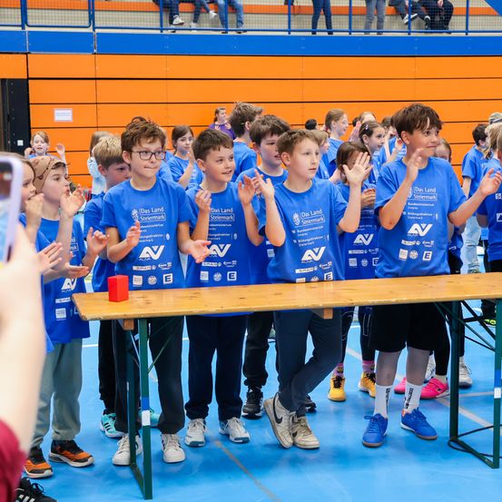 Eine Gruppe junger Jungen in blauen T-Shirts steht hinter einem Tisch bei einer Veranstaltung, klatscht und lächelt. Ein roter Kasten liegt auf dem Tisch. Dahinter sind weitere Kinder und Erwachsene anwesend.
