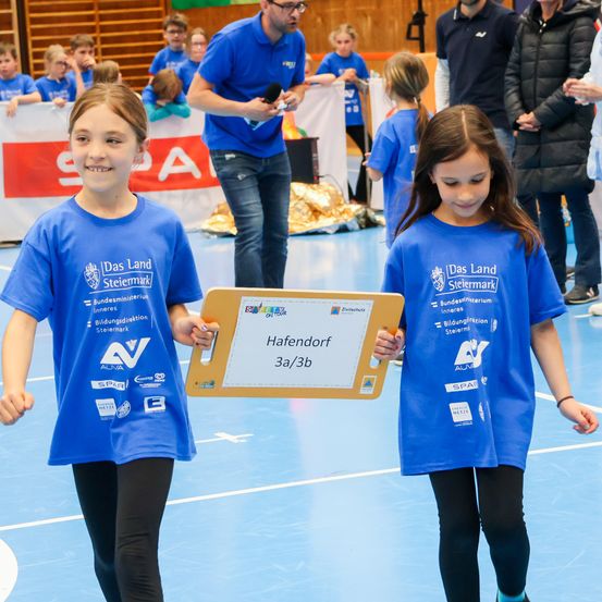 Zwei Mädchen halten ein Zertifikat für den 3. Platz bei einer Veranstaltung in den Händen und tragen passende blaue T-Shirts mit Logos. Dahinter spricht ein Mann mit Brille in ein Mikrofon. Zuschauer in unterschiedlicher Kleidung beobachten im Hintergrund.