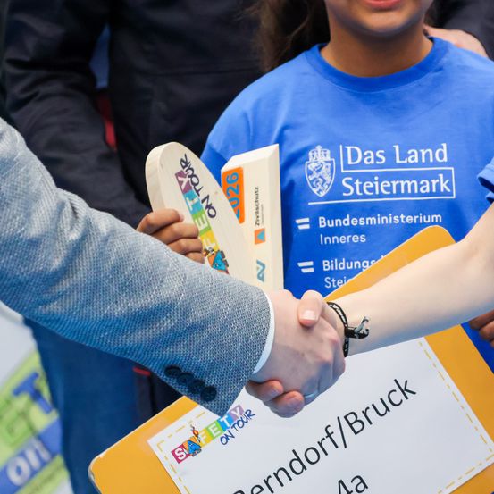 Eine Person in einem Anzug gibt einem jungen Jungen in einem blauen T-Shirt mit 'Das Land Steiermark' die Hand. Der Junge hält ein Buch mit 'Sicherheit auf Tour'. Eine dritte Person steht hinter ihnen.