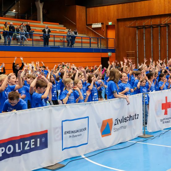 Eine Gruppe von Kindern in blauen T-Shirts jubelt in einer Indoor-Sportanlage mit Bannern und Holzwänden. Einige Personen stehen im Hintergrund auf Tribünen.