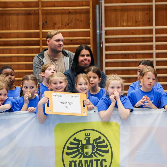 Eine Gruppe von Kindern in blauen T-Shirts, einige mit Snacks, schauen nach vorne. Ein Mädchen hält eine gelbe Karte. Dahinter eine Holzwand und Erwachsene.