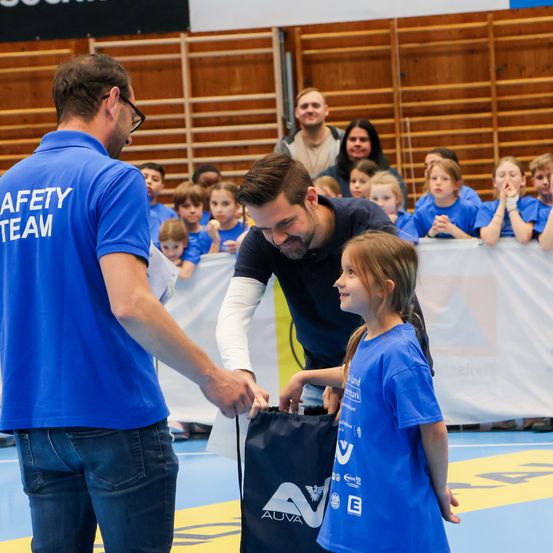 Ein Mann in einem schwarzen Shirt übergibt einer jungen Mädchen in einer Turnhalle einen blauen Beutel, während ein Mann in einem blauen Sicherheitsteam-Shirt zuschaut. Dahinter beobachten mehrere Kinder.