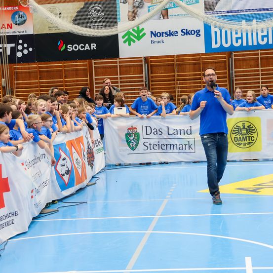 Ein Mann spricht in ein Mikrofon auf einem Sportplatz, umgeben von Kindern in blauen Shirts. Werbung für verschiedene Organisationen hängt im Hintergrund.