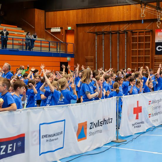 Eine Gruppe von Kindern in blauen T-Shirts mit dem Zivilschutz-Logo jubelt bei einer Sportveranstaltung. Hinter ihnen sind Banner zu sehen. Im Hintergrund sind eine Holzwand, Tribünen und eine Treppe sichtbar.