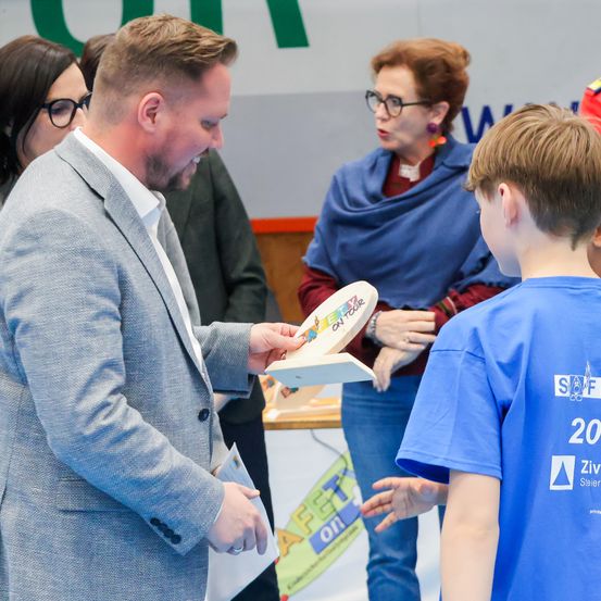 Ein Mann in einem grauen Anzug übergibt einem Jungen in einem blauen T-Shirt ein Buch. Dahinter beobachtet eine ältere Frau mit Brille und blauer Schürze. Ein farbenfrohes Poster mit Buchstaben ist im Hintergrund.