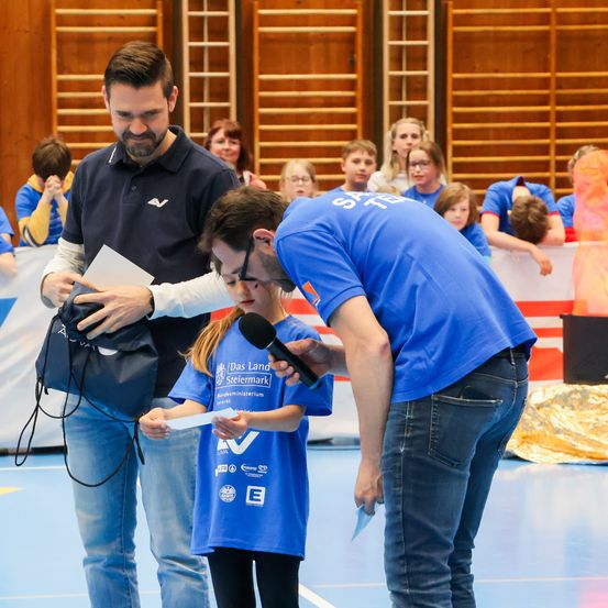 Ein junges Mädchen in einem blauen Shirt hält ein Papier und ein Mikrofon, während zwei Männer über ihre Schulter schauen. Das Event findet in einer Sporthalle statt, mit Zuschauern und hölzernen Tribünen im Hintergrund.