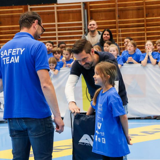 Ein Mann in einem blauen T-Shirt mit der Aufschrift 'Sicherheitsteam' überreicht einer kleinen Mädchen in einem blauen T-Shirt ein Geschenk. Die Veranstaltung findet in einer Turnhalle statt, mit mehreren Personen in blauen T-Shirts im Hintergrund.