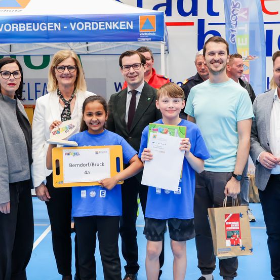 Eine Gruppe von Menschen steht in einer Halle mit einem Banner. Zwei Kinder halten Urkunden und Taschen. Die Frau lächelt mit der Hand auf dem Kopf des Mädchens.