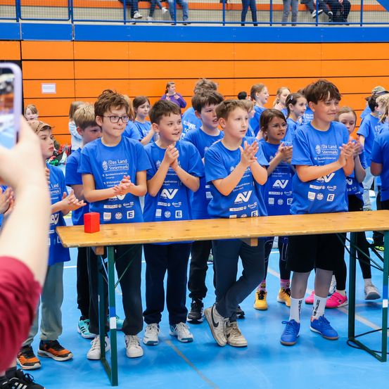 Mehrere junge Jungen in blauen T-Shirts stehen an einem Tisch und klatschen in die Hände. Der Junge in der Mitte hält einen roten Würfel. Sie scheinen sich in einer Sportveranstaltung zu befinden.