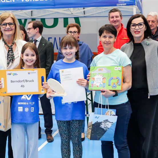 Zwei Mädchen stehen vor einem Eventzelt mit Erwachsenen, halten ein Zertifikat und ein Buch in den Händen und posieren lächelnd für ein Foto.