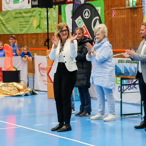 Eine Gruppe von Menschen steht auf einem Sportplatz und klatscht. Eine Frau in Weiß führt, neben einer Frau in Blau. Hinter ihnen zeigt ein Banner Text und Logos. Kinder sitzen auf einer Bank im Hintergrund.