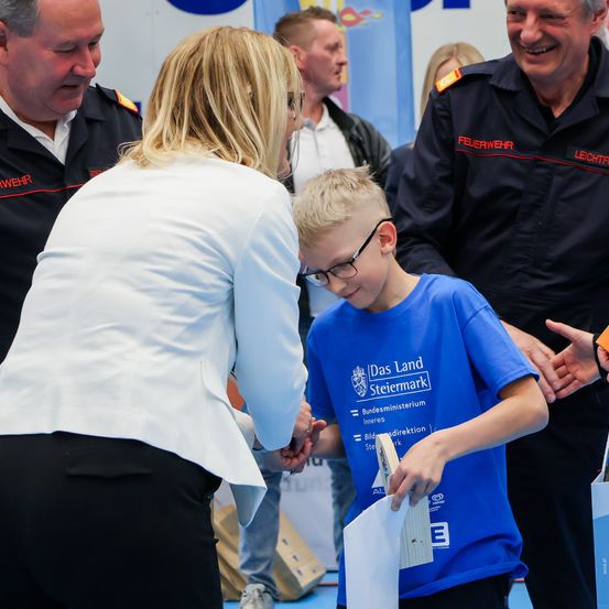 Ein junger Junge mit Brille hält ein Papier in der Hand und erhält von einer Frau einen Preis. Hinter ihnen stehen mehrere Menschen, darunter ein Mann in einer Feuerwehruniform.