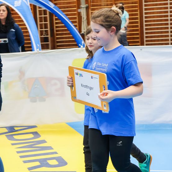 Zwei Mädchen halten ein Schild mit der Aufschrift 'Knottinger 4a' und gehen auf einem Spielfeld. Eine weitere Mädchen steht hinter ihnen, und eine Frau ist im Hintergrund.