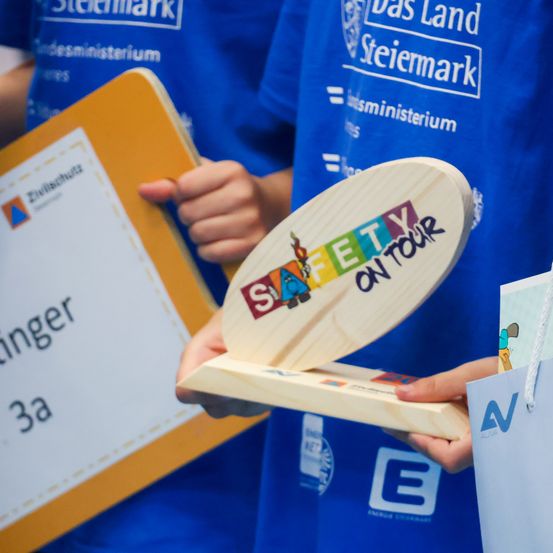 Eine Person hält eine hölzerne Plakette, auf der Safety on Tour steht. Daneben befindet sich ein gelber Ordner mit der Aufschrift Zivilschutz.