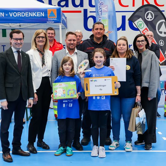 Eine Gruppe von Menschen steht auf einem blauen Boden, wahrscheinlich drinnen, mit zwei Mädchen, die ein Buch und ein Zertifikat halten. Ein Zelt mit Text und einer Flagge ist hinter ihnen. Sie scheinen bei einer Veranstaltung zu sein.