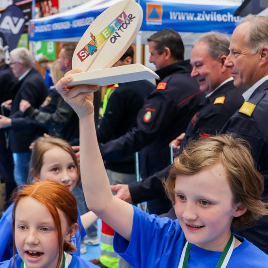 Zwei Kinder halten ein Holzschild mit 'Sicherheit auf Tour' hoch, während eine Menge Menschen zuschaut.
