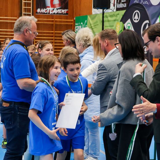 Eine Gruppe junger Kinder, die blaue Uniformen und Medaillen tragen, steht in einem Kreis mit Erwachsenen, möglicherweise in einer Schulumgebung. Sie halten Zertifikate und scheinen eine Leistung zu feiern.
