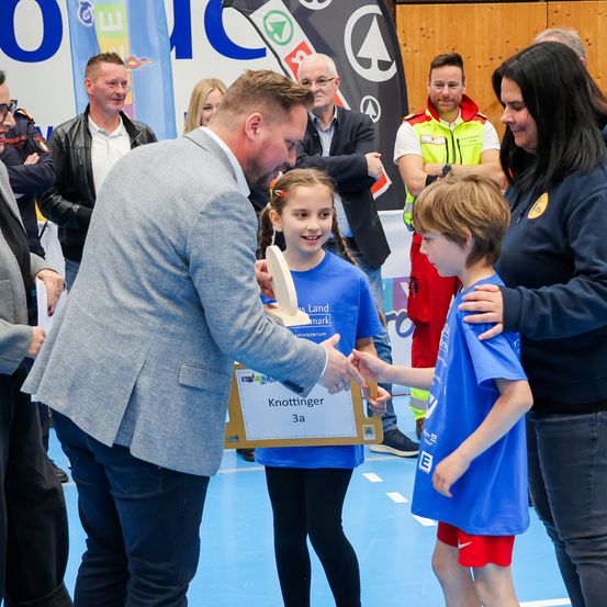 Ein Mann überreicht einem Jungen eine Trophäe. Ein Mädchen und eine Frau stehen in der Nähe. Dahinter ist ein Banner mit Logos und Text zu sehen. Mehrere Erwachsene stehen im Hintergrund und beobachten.