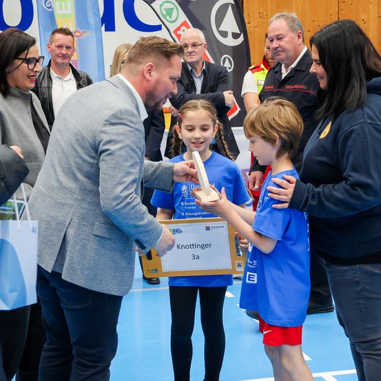 Ein Mann in einem grauen Anzug überreicht einem jungen Mädchen ein Zertifikat und eine Schachtel. Ein Junge in Blau hält die Schachtel. Sie werden von Erwachsenen in einer Turnhalle umgeben.