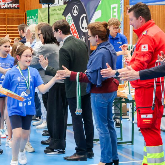 Ein junges Mädchen in einem blauen Shirt erhält eine Medaille von einer Frau in einer roten Jacke. Sie werden von einer Gruppe von Erwachsenen in einer Halle umgeben.