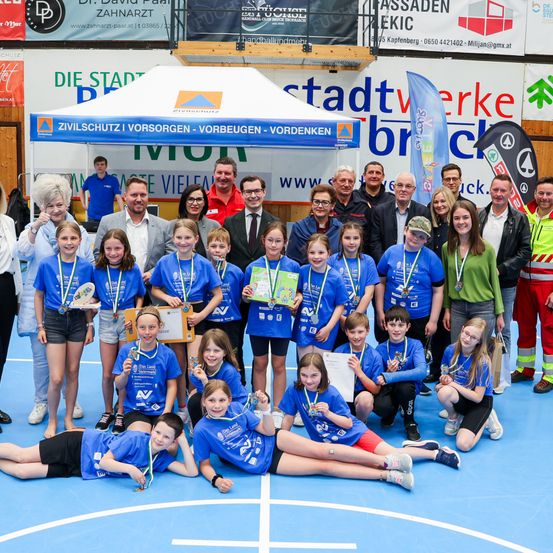 Eine Gruppe von Kindern in blauen Shirts posiert für ein Foto mit Erwachsenen unter einem Zelt, während sie Medaillen und Urkunden halten. Es scheint sich um eine Preisverleihung zu handeln.