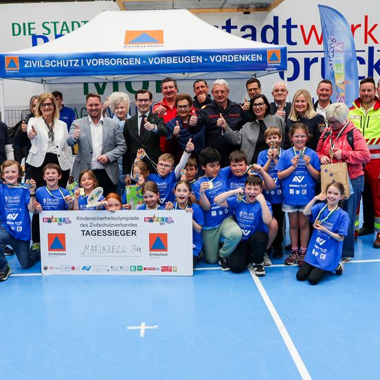 Gruppenfoto der Gewinner eines Sicherheitswettbewerbs unter einem Zelt mit einem Banner. Kinder und Erwachsene, alle in blauen Shirts, stehen und sitzen auf einem blauen Boden. Sie halten Medaillen und einige haben Brillen.