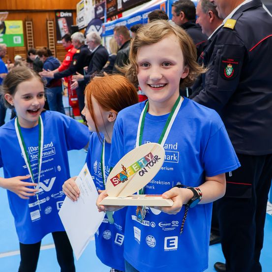 Drei Kinder in blauen T-Shirts halten eine Holzplakette und lächeln, während eine Menschenmenge im Hintergrund steht. Eine Person in Uniform steht in der Nähe.