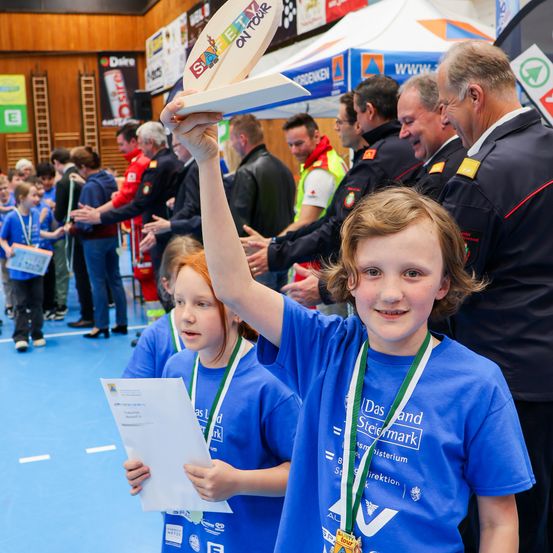 Eine Gruppe von Kindern in blauen T-Shirts und mit Medaillen steht lächelnd vor einer Menschenmenge. Ein junges Mädchen hält ein Zertifikat und eine Medaille.