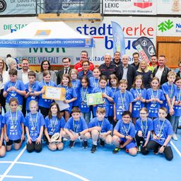 Eine Gruppe junger Athleten in blauen Shirts und mit Medaillen posiert für ein Foto mit Erwachsenen, einige halten Urkunden, bei einer Sportveranstaltung.