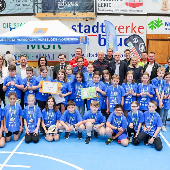 Eine Gruppe junger Athleten in blauen Shirts und mit Medaillen posiert für ein Foto mit Erwachsenen, einige halten Urkunden, bei einer Sportveranstaltung.