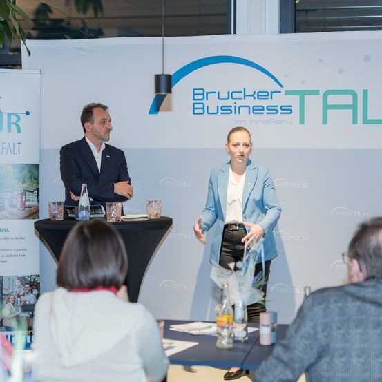 Zwei Fachleute stehen an einem Podium mit einem Hintergrund von Bruckner Business TAL. Eine Frau in einem blauen Blazer präsentiert, während ein Mann im Anzug zuhört. Zuhörer sitzen um einen Tisch vor ihnen.