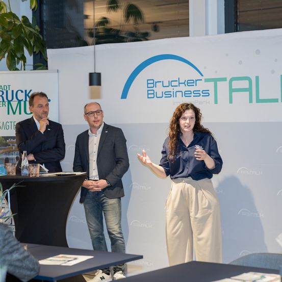 Drei Personen stehen vor einem weißen Banner mit 'Brucker Business Park'. Eine Frau spricht und gestikuliert, während zwei Männer zuhören. Tische und Stühle sind in der Nähe aufgebaut.