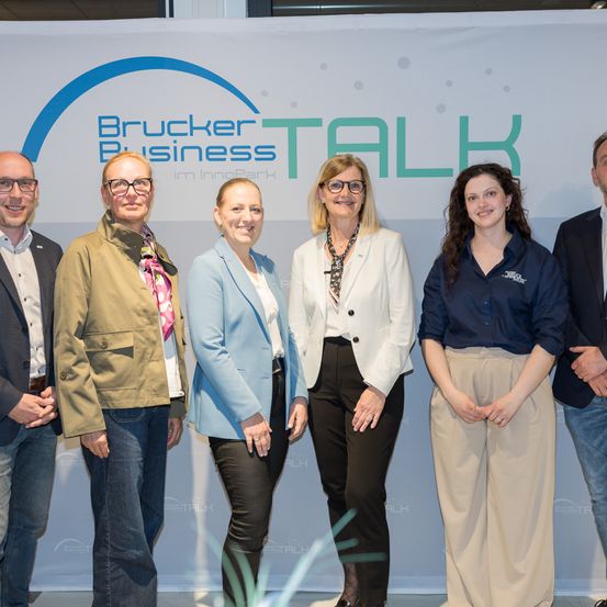 Sechs Personen, alle schick gekleidet, stehen vor einem Banner mit den Worten Brucker Business Talk. Das Banner hat einen blauen Kreis und Punkte. Die Leute lächeln für ein Foto.