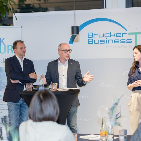 Drei Personen stehen hinter einem Tisch mit dem Schild Bruckner Business. Zwei Männer und eine Frau, alle schick gekleidet, sprechen zu einem Publikum.