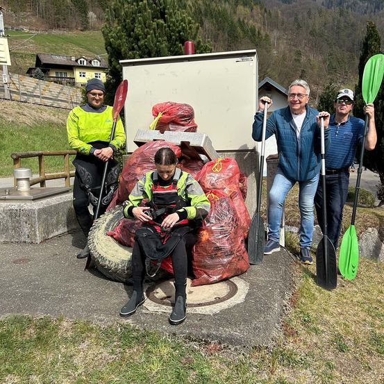 Vier Personen stehen neben einem Müllhaufen und halten Paddel in der Hand. Eine Frau sitzt auf einem Reifen mit Müllsäcken in der Nähe. Hinter ihnen stehen Häuser und Berge.