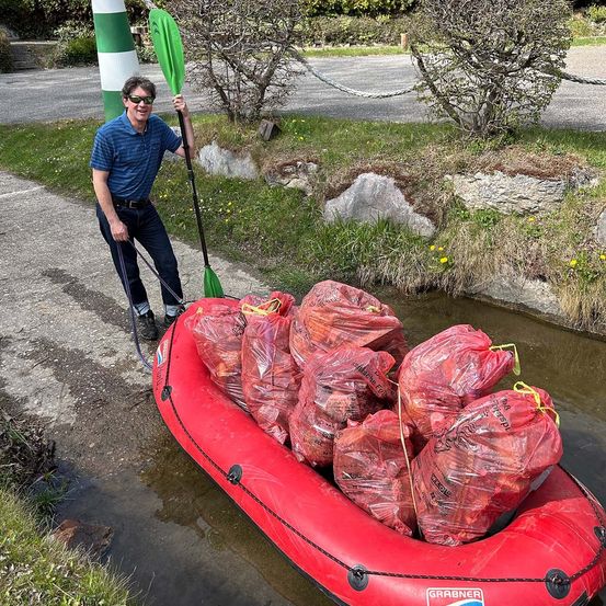 Ein Mann mit Sonnenbrille zieht einen roten Schlauchboot voller roter Beutel mit einem grünen Paddel auf einem Bach.