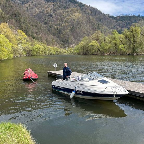 Ein Mann steht neben seinem Boot am Steg, ein rotes aufblasbares Boot schwimmt in der Nähe auf einem ruhigen See mit Bergen im Hintergrund.