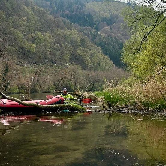 Eine Person in einer grünen Jacke paddelt mit einem roten Schlauchboot auf einem Fluss, umgeben von grünen Bäumen und Büschen.